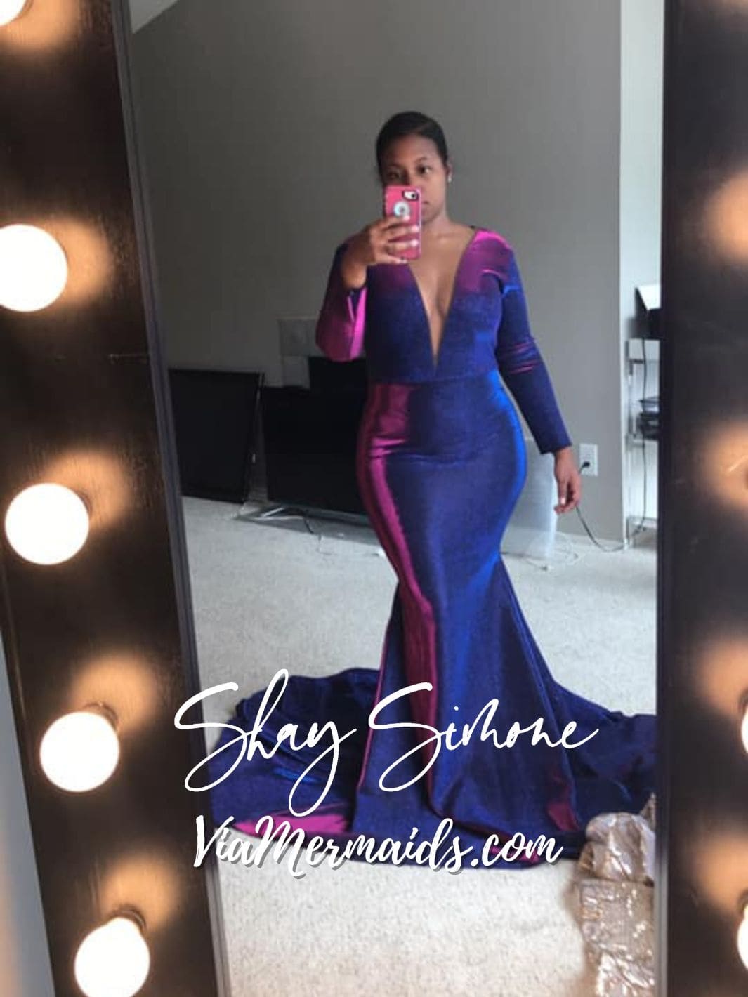 A woman takes a selfie in a lighted mirror while wearing a stunning, long-sleeved, mermaid-style gown with plunging neckline in shifting shades of blue and pink.