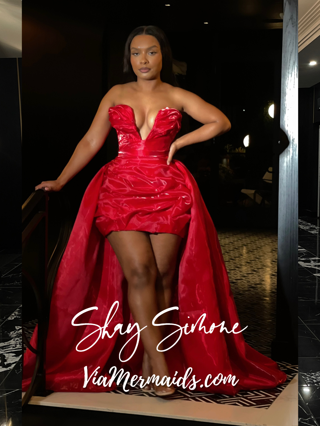 A woman models a strapless, shiny, bright red corset dress with a structured bodice, deep neckline, and voluminous skirt in a hotel lobby. The dress also features a detachable train.