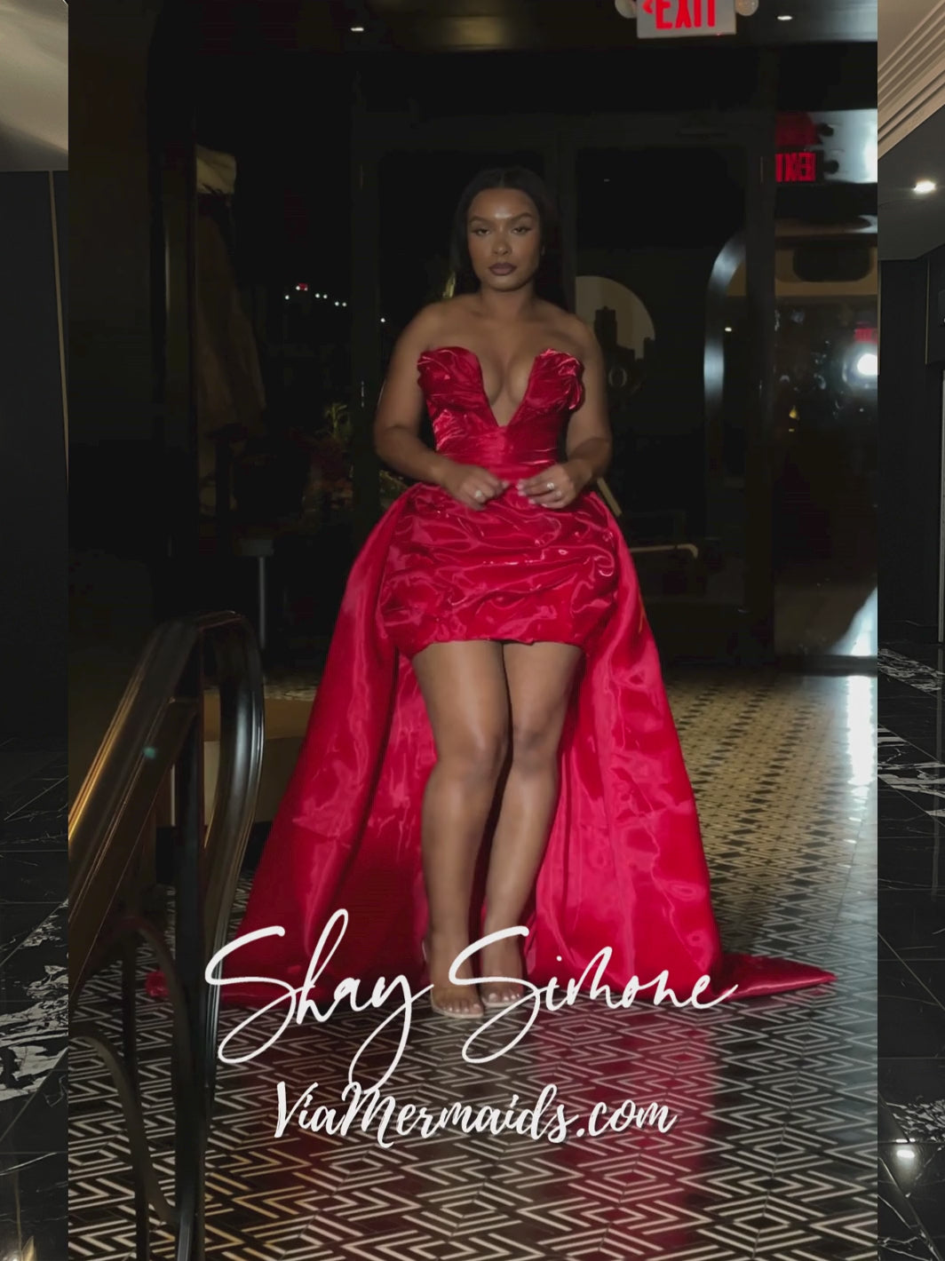 A video of a woman modelling a strapless, shiny, bright red corset dress with a structured bodice, deep neckline, and voluminous overskirt in a hotel lobby.